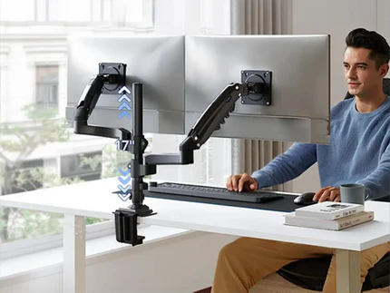 A man sits at a desk working on two computer monitors mounted on adjustable arms.