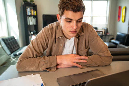 A young man sits at a table, looking intently at his laptop screen with a focused expression.