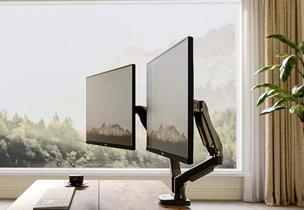 A desk with dual monitors showing a mountain scene, near a window with a scenic view and a plant.